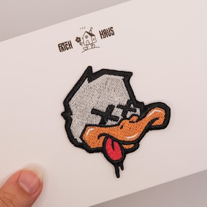 Puede incluir: Parche bordado con la cabeza de un pato de dibujos animados con plumas grises, pico naranja y lengua roja. Los ojos están tachados con líneas negras. El parche tiene un borde negro sobre un fondo blanco.