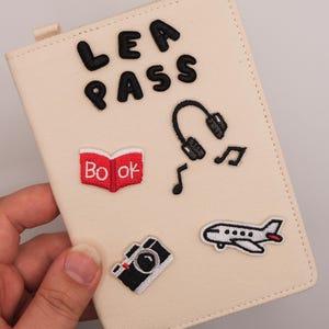 Könnte beinhalten: Cremefarbener Reisepasshalter mit aufgestickten Patches zum Thema Reisen. Mit den Worten "LEA PASS", einem Buch, Kopfhörern, Noten, einer Kamera und einem Flugzeug. Der Halter ist rechteckig mit einer kleinen Lasche.