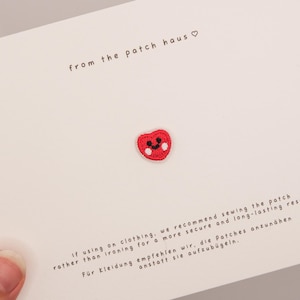 Può includere: Una toppa a forma di cuore rosso con un volto sorridente e guance bianche, esposta su una carta bianca. Il testo "from the patch haus" è sopra la toppa. Il testo sottostante consiglia di cucire la toppa.