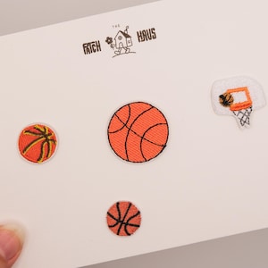 Könnte beinhalten: Eine weiße Karte mit gestickten Basketball-Patches. Die Patches zeigen drei Basketbälle in verschiedenen Größen und einen Basketballkorb mit Netz. Der Text "The Frich Haus" ist oben aufgedruckt.