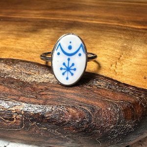 May include: Oval-shaped ring with a white ceramic face featuring blue geometric designs. The ring has a silver-colored band. The ring is displayed on a wooden surface with a natural wood grain.