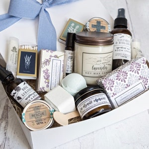 K&ouml;nnte beinhalten: Eine wei&szlig;e Geschenkbox, gebunden mit einer blauen Schleife, gef&uuml;llt mit Pflegeprodukten. Artikel umfassen eine Lavendel-Sojawachskerze, &auml;therisches &Ouml;lspray, Badebomben, Lippenbalsam, Seife und einen Haut-Rettungsbalsam. Die Box ist mit Stroh gef&uuml;llt.