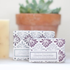 May include: Two bars of white handmade soap with lavender and lemongrass scent. The soap is packaged in a box with a purple and gray floral pattern. The text on the box reads "Little Flower Handmade Soap Lavender Lemongrass Relaxing, Fresh Scent Lemongrass Soap".