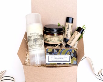 May include: A brown cardboard box with a lid open, revealing a gift set of men's grooming products. The set includes a natural deodorant stick, a small jar of beard balm, a bar of soap, and two lip balms. The box is filled with shredded paper and has a black and white ribbon tied around it.