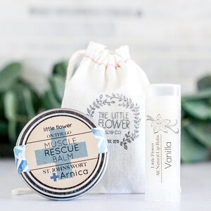 May include: A set of natural skincare products, including a round tin of Muscle Rescue Balm, a vanilla lip balm, and a small drawstring bag. The balm tin has a blue and white label with the words "Muscle Rescue Balm". The lip balm is white with a vanilla flower design.
