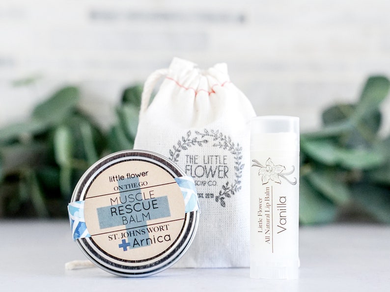 May include: A set of natural skincare products, including a round tin of Muscle Rescue Balm, a vanilla lip balm, and a small drawstring bag. The balm tin has a blue and white label with the words "Muscle Rescue Balm". The lip balm is white with a vanilla flower design.