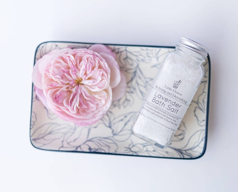 Op de afbeelding: Een rechthoekige keramische schaal met een bloemenpatroon bevat een grote roze roos. Een heldere glazen fles Lavendel Badzout staat naast de roos. De fles heeft een zilveren deksel en een wit etiket.