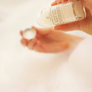 Op de afbeelding: Een heldere glazen fles Lavendel Badzout wordt boven een bad gehouden. Op het etiket staat "Little Flower Relaxing and Detoxifying Lavender Bath Salt". De fles wordt in het badwater gegoten. Het badwater is wit en schuimig.