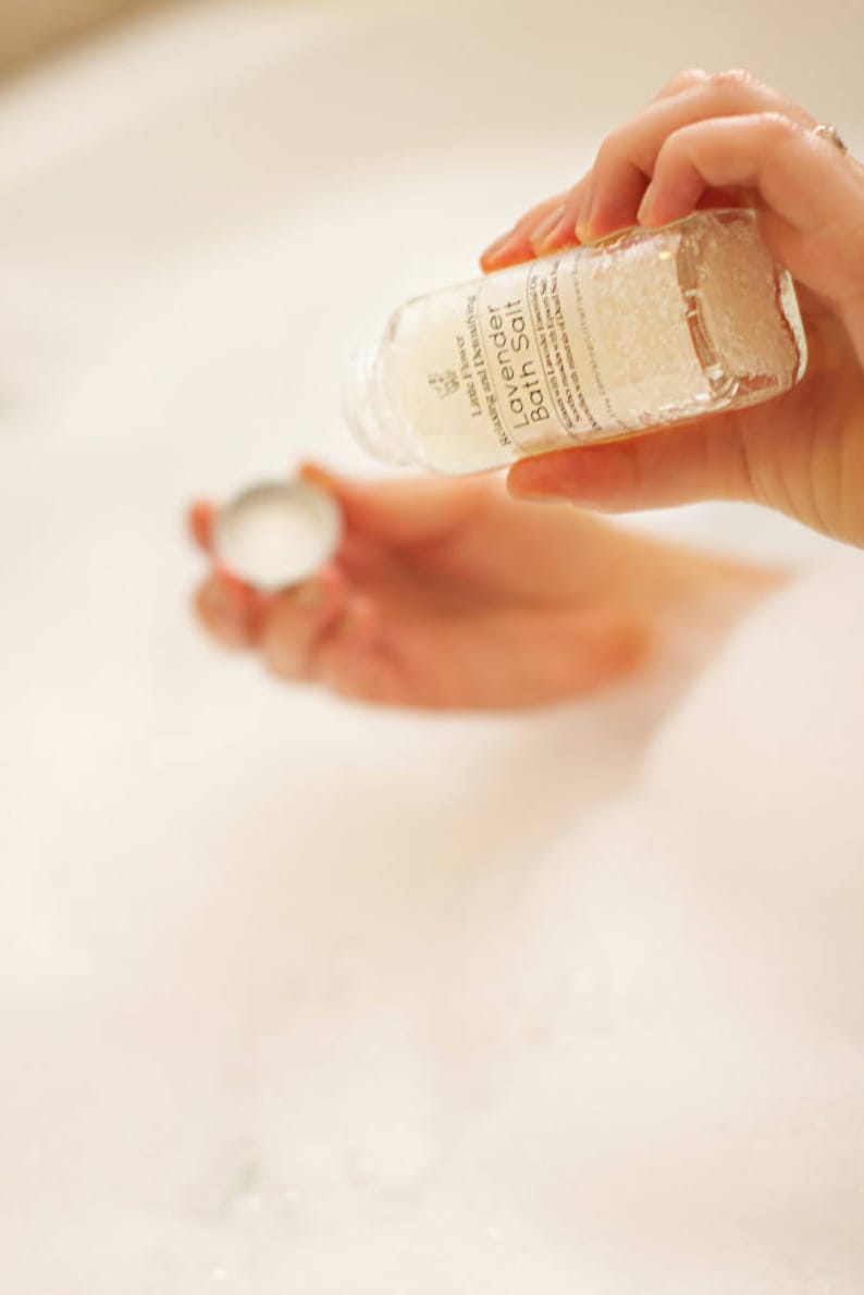 Op de afbeelding: Een heldere glazen fles Lavendel Badzout wordt boven een bad gehouden. Op het etiket staat "Little Flower Relaxing and Detoxifying Lavender Bath Salt". De fles wordt in het badwater gegoten. Het badwater is wit en schuimig.