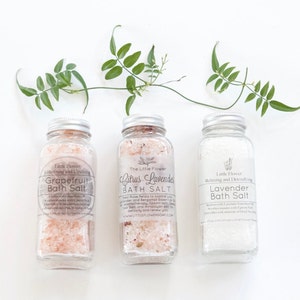 Op de afbeelding: Drie glazen potten met zilveren deksels, gevuld met badzouten. De etiketten luiden "Grapefruit", "Citrus-Lavendel" en "Lavendel". De potten bevatten roze, roze-gespikkelde en witte zouten. Groene plantentakken zijn erboven geplaatst.
