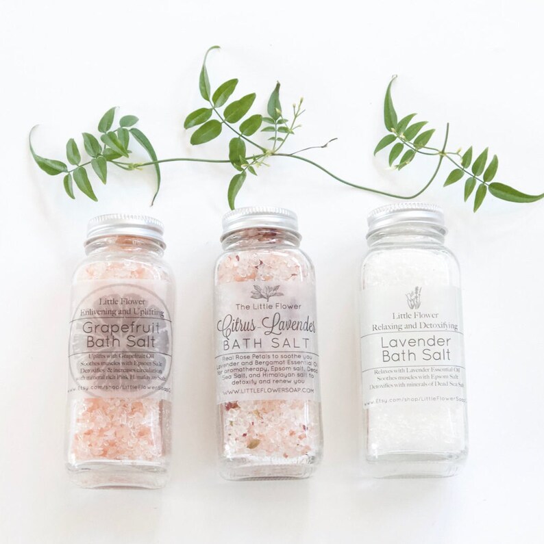 Op de afbeelding: Drie glazen potten met zilveren deksels, gevuld met badzouten. De etiketten luiden "Grapefruit", "Citrus-Lavendel" en "Lavendel". De potten bevatten roze, roze-gespikkelde en witte zouten. Groene plantentakken zijn erboven geplaatst.