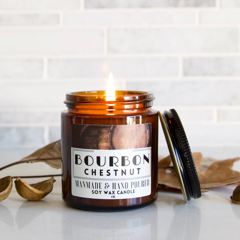 Puede incluir: Un tarro de vela de vidrio marr&oacute;n con una etiqueta blanca que dice "Bourbon Chestnut" y "Handmade & Hand Poured Soy Wax Candle 4 oz". La vela est&aacute; encendida y la llama es visible.