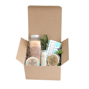 May include: A brown cardboard box filled with a spa gift set. The set includes a jar of grapefruit bath salt, a tube of lip balm, a small jar of rescue salve, a bar of tea tree and lime soap, and a sprig of rosemary.