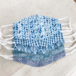 May include: Set of five blue and white patterned face masks with white elastic ear loops. The masks feature different geometric patterns, including chevron, gingham, and floral designs.