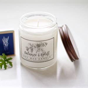 May include: A white soy wax candle in a clear glass jar with a brown lid. The label reads "Balance & Uplift Soy Wax Candle Eucalyptus,  Eucalyptus & Lavender".
