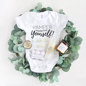 May include: A white baby onesie with the text "Pamper Yourself!" is surrounded by eucalyptus leaves. The image includes a small tin, several lavender-scented items, a bottle of baby bump body butter, and a jar of soy wax candle.