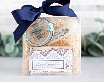 May include: A gift set in a clear box tied with a navy blue ribbon. The set includes a tin of "Skin Rescue Balm", a "Raspberry" lip balm, and a bar of "Lavender Lemongrass" soap. The soap is labeled "Relaxing, Fresh Scent, Everyday Soap".