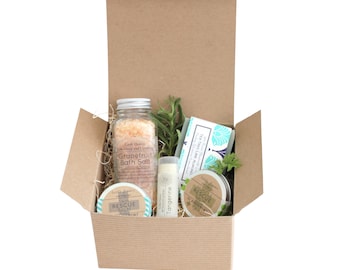May include: A brown cardboard box with a lid open, revealing a gift set of bath and body products. The set includes a jar of grapefruit bath salt, a tube of tangerine lip balm, a small jar of rescue balm, a bar of tea tree and lime almond soap, and a sprig of rosemary.