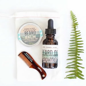 May include: A beard grooming kit with a balm, oil, and comb. The balm is in a silver tin with a teal and white label. The oil is in a brown glass bottle with a dropper. A brown comb and a sprig of green fern complete the set.