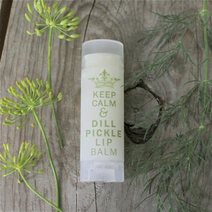 May include: A white tube of lip balm with a green label that reads "Keep Calm & Dill Pickle Lip Balm". The label also features a crown graphic. The lip balm is sitting on a wooden surface with green dill sprigs.
