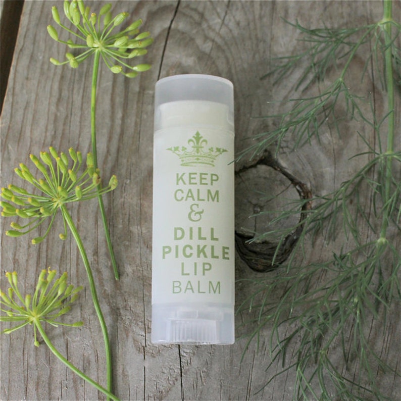 May include: A white tube of lip balm with a green label that reads "Keep Calm & Dill Pickle Lip Balm". The label also features a crown graphic. The lip balm is sitting on a wooden surface with green dill sprigs.