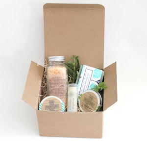 May include: A brown cardboard box filled with a spa gift set. The set includes a jar of grapefruit bath salt, a small jar of muscle rescue balm, a tube of tangerine lip balm, a bar of tea tree lime almond soap, and a sprig of rosemary.