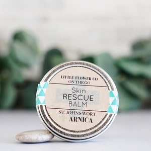 May include: A small, round metal tin of Skin Rescue Balm. The label features the text "LITTLE FLOWER CO. ON THE GO" and "Skin RESCUE BALM". The label also includes "ST. JOHNS WORT ARNICA". The tin is accented with a teal and white geometric pattern.