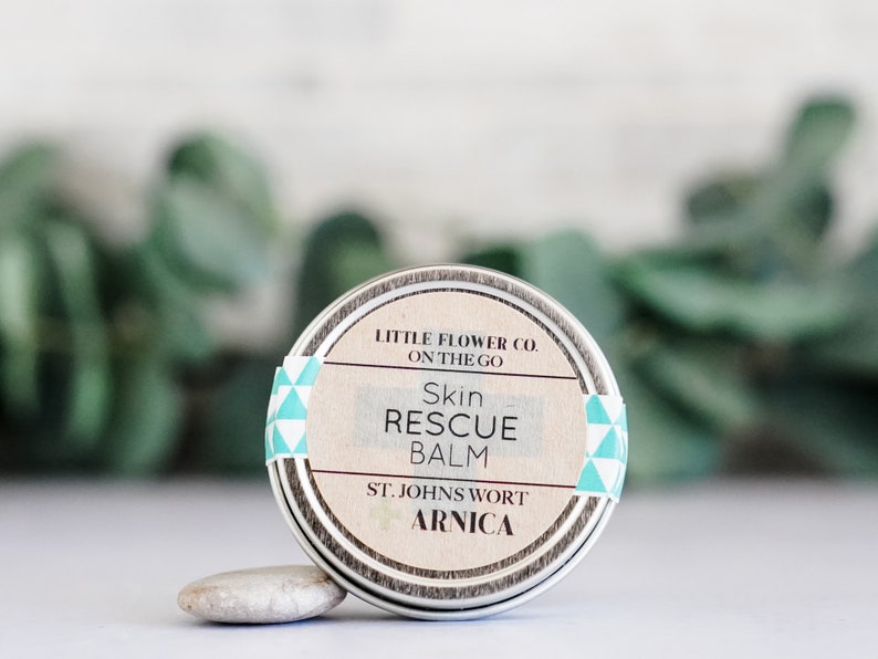 May include: A small, round metal tin of Skin Rescue Balm. The label features the text "LITTLE FLOWER CO. ON THE GO" and "Skin RESCUE BALM". The label also includes "ST. JOHNS WORT ARNICA". The tin is accented with a teal and white geometric pattern.