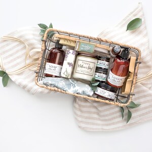 Puede incluir: Una cesta de regalo llena de productos de cuidado personal, incluyendo jab&oacute;n, loci&oacute;n, una vela y exfoliantes. La cesta es de material tejido y se encuentra sobre una tela a rayas con hojas verdes. La vela dice "Happy Mother's Day!"