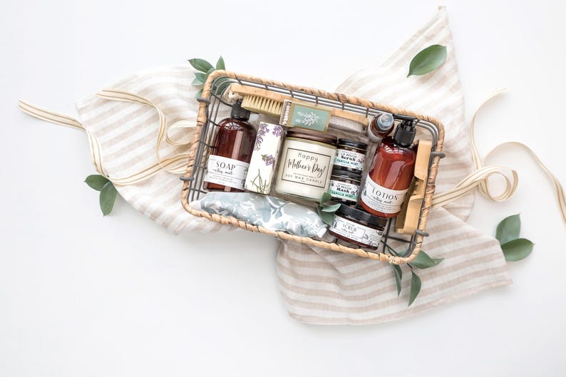 Puede incluir: Una cesta de regalo llena de productos de cuidado personal, incluyendo jab&oacute;n, loci&oacute;n, una vela y exfoliantes. La cesta es de material tejido y se encuentra sobre una tela a rayas con hojas verdes. La vela dice "Happy Mother's Day!"