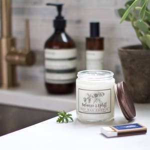 May include: A white soy wax candle with a brown lid and a label that reads "Balance & Uplift". The candle is sitting on a white countertop next to a matchbox and a sprig of green foliage.