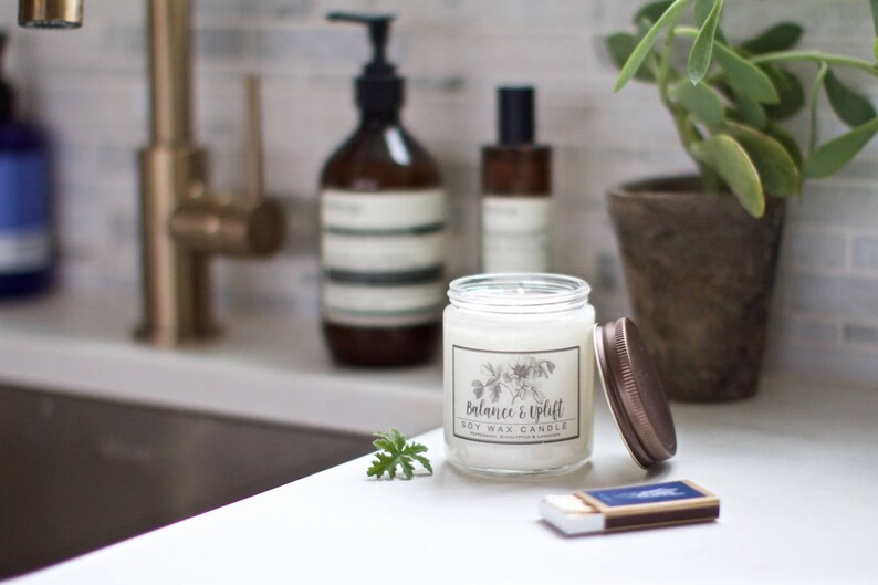 May include: A white soy wax candle with a brown lid and a label that reads "Balance & Uplift". The candle is sitting on a white countertop next to a matchbox and a sprig of green foliage.