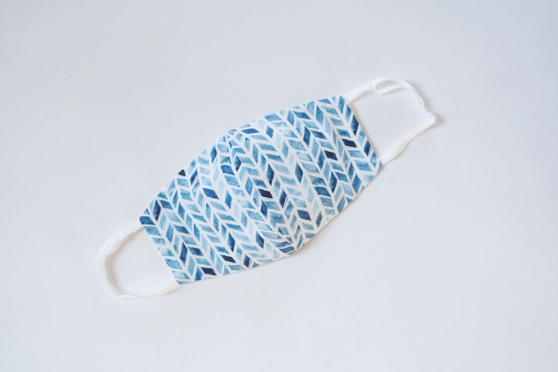 May include: A white face mask with blue and white geometric pattern. The mask has white elastic straps.