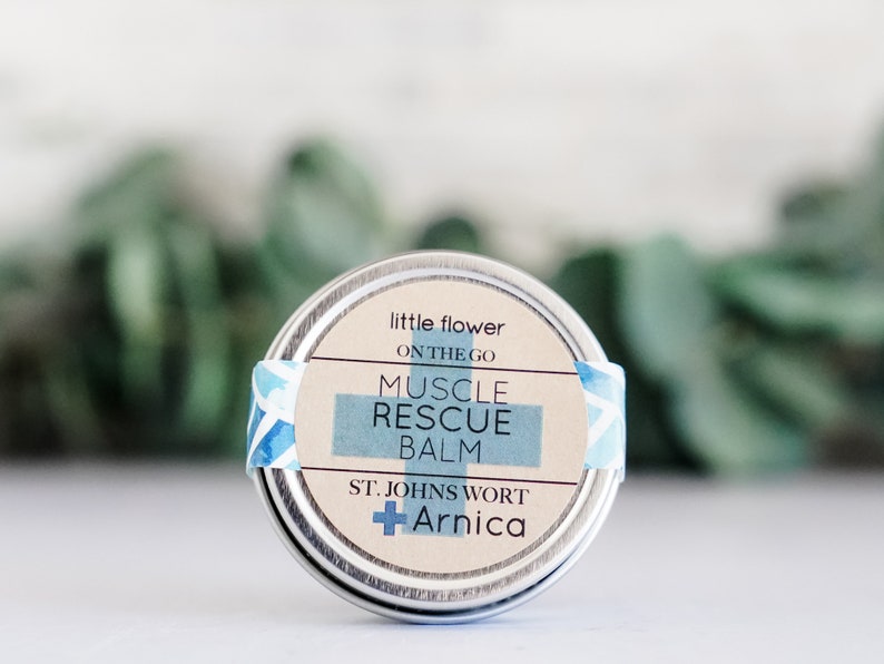 May include: A small, round metal tin of Muscle Rescue Balm. The label features the text "little flower on the go" and "Muscle Rescue Balm" in a light blue rectangle. The label also includes "St. John's Wort + Arnica".