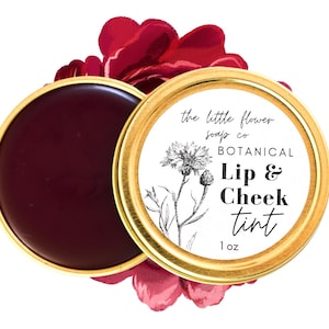 May include: A gold tin with a white label that reads "the little flower soap co BOTANICAL Lip & Cheek Tint 1 oz". The tin contains a red lip and cheek tint.