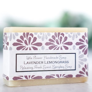 May include: A bar of handmade soap with a white label that reads "Little Flower Handmade Soap Lavender Lemongrass Relaxing, Fresh Scent, Everyday Soap". The soap is wrapped in a white paper with a floral pattern in shades of purple and gray.