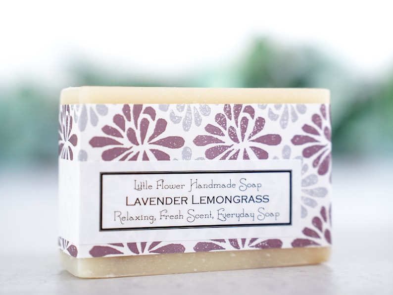 May include: A bar of handmade soap with a white label that reads "Little Flower Handmade Soap Lavender Lemongrass Relaxing, Fresh Scent, Everyday Soap". The soap is wrapped in a white paper with a floral pattern in shades of purple and gray.