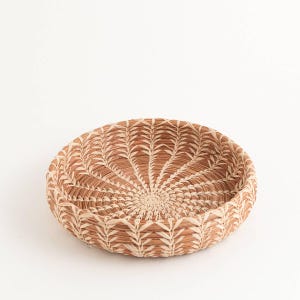 May include: A round, woven basket with a natural, earthy color palette. The basket features a detailed pattern radiating from the center, with alternating light and dark brown tones. The design is reminiscent of a sunburst or floral motif.