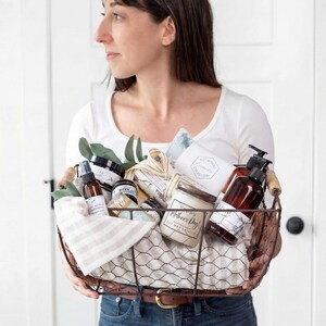 Puede incluir: Una cesta de alambre llena de productos de cuidado personal, incluyendo una vela, loci&oacute;n y botellas de spray. La cesta est&aacute; forrada con una tela a rayas y vegetaci&oacute;n. Los productos est&aacute;n en envases marrones y blancos. La cesta tiene un asa de madera.