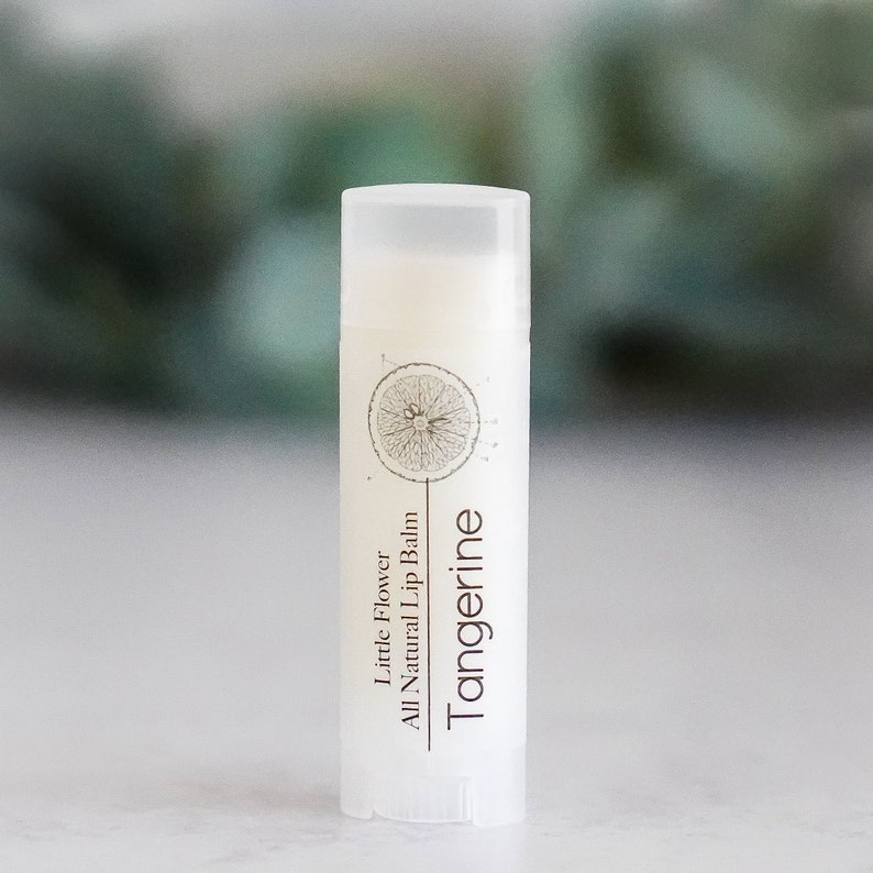Puede incluir: Un tubo blanco de b&aacute;lsamo labial con aroma a mandarina con una ilustraci&oacute;n en blanco y negro de una rodaja de mandarina en la etiqueta. La etiqueta tambi&eacute;n dice "Little Flower All Natural Lip Balm Tangerine".