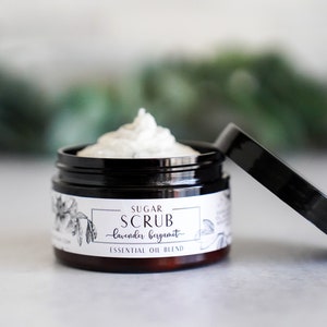 May include: A black jar with a white label that reads "Sugar Scrub - Lavender Bergamot - Essential Oil Blend". The jar is open and filled with a white, creamy scrub.