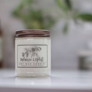 May include: A clear glass candle jar with a copper lid. The label reads "Balance & Uplift Soy Wax Candle" with a floral design.