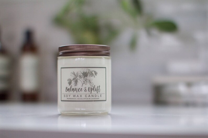 May include: A clear glass candle jar with a copper lid. The label reads "Balance & Uplift Soy Wax Candle" with a floral design.