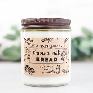 May include: A brown glass candle jar with a brown lid and a kraft paper label. The label reads "Little Flower Soap Co. Handmade Banana Nut Bread Soy Wax 8 oz.". The label has a black and white illustration of baking ingredients and tools.