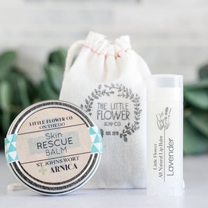May include: A skincare set featuring a round tin of Skin Rescue Balm, a tube of Lavender lip balm, and a small drawstring bag. The balm tin has a label with text, and the lip balm tube has a lavender illustration. The bag has a floral design.