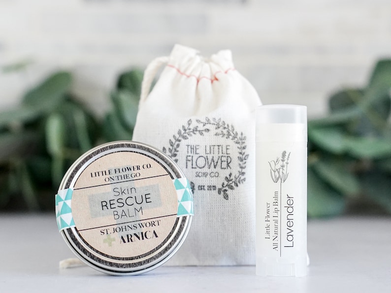 May include: A skincare set featuring a round tin of Skin Rescue Balm, a tube of Lavender lip balm, and a small drawstring bag. The balm tin has a label with text, and the lip balm tube has a lavender illustration. The bag has a floral design.