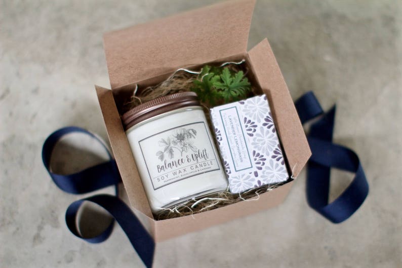 May include: A brown cardboard box with a lid open, revealing a white soy wax candle with a brown lid and a label that reads "Balance & Uplift Soy Wax Candle". The candle is surrounded by brown shredded paper and a small green plant. A white bar of soap with a purple and white floral pattern is also in the box.