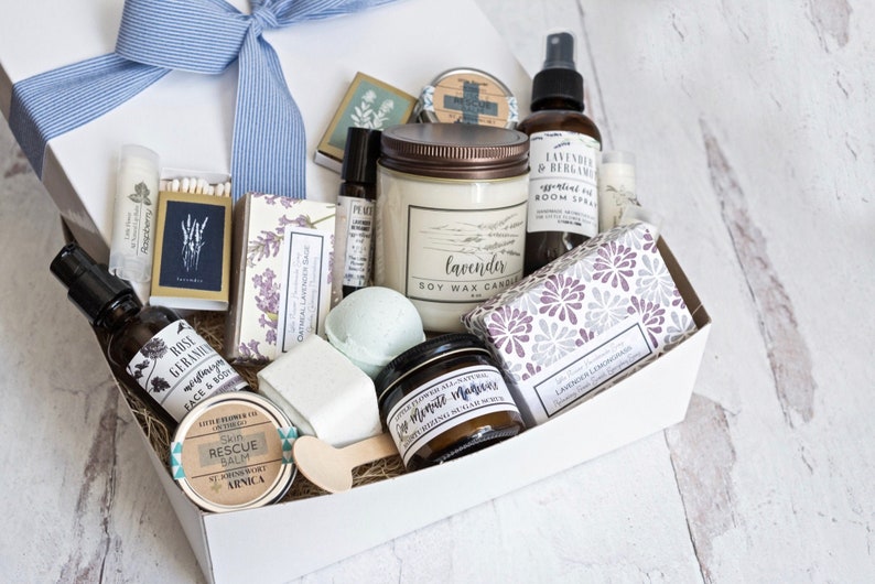 K&ouml;nnte beinhalten: Eine wei&szlig;e Geschenkbox, gebunden mit einer blauen Schleife, gef&uuml;llt mit Pflegeprodukten. Artikel umfassen eine Lavendel-Sojawachskerze, &auml;therisches &Ouml;lspray, Badebomben, Lippenbalsam, Seife und einen Haut-Rettungsbalsam. Die Box ist mit Stroh gef&uuml;llt.