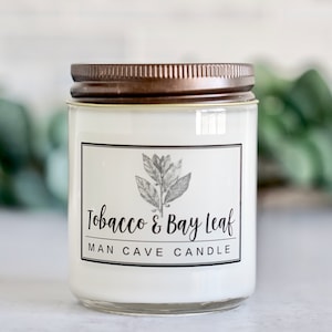 May include: A clear glass candle jar with a brown lid. The label reads "Tobacco & Bay Leaf Man Cave Candle" and features a black and white illustration of a bay leaf.