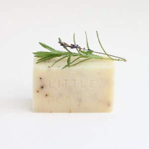 May include: A white bar of soap with the words "LITTLE FLOWER" embossed on it. A sprig of lavender is resting on top of the soap.